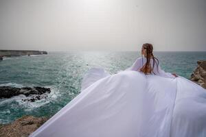 glücklich Freiheit Frau auf das Strand genießen und posieren im Weiß Kleid. Rückseite Aussicht von ein Mädchen im ein flattern Weiß Kleid im das Wind. Feiertage, Ferien beim Meer. foto