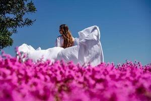 ein schön Frau im ein Weiß fließend lange Kleid bleibe in der Nähe von ein schön Feld mit Rosa Blumen Rückseite Aussicht foto