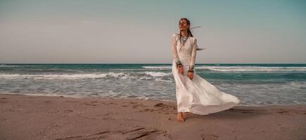 Modell- im Boho Stil im ein Weiß lange Kleid und Silber Schmuck auf das Strand. ihr Haar ist geflochten, und Dort sind viele Armbänder auf ihr Waffen. foto