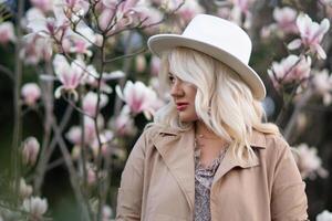 ein blond Frau tragen ein Weiß Hut steht im Vorderseite von ein Baum mit Rosa Blumen foto