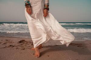 Modell- im Boho Stil im ein Weiß lange Kleid und Silber Schmuck auf das Strand. ihr Haar ist geflochten, und Dort sind viele Armbänder auf ihr Waffen. foto