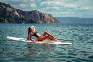 Frau Saft Meer. Sport Mädchen auf ein Surfbrett im das Meer auf ein sonnig Sommer- Tag. im ein schwarz Baden Anzug, er sitzt auf ein Sapa im das Meer. sich ausruhen auf das Meer. foto