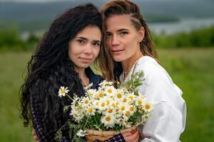 zwei Frauen genießen Natur im ein Feld von Gänseblümchen. Freundinnen umarmen halt ein Strauß von Gänseblümchen und aussehen beim das Kamera. foto