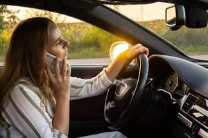 jung Frau mit Handy, Mobiltelefon Telefon während Fahren Auto auf Autobahn Straße während Sonnenuntergang. Fahrerin hat Unfall Berufung mit Smartphone zum Hilfe. Geschäft Frau beschäftigt Fahren Konzept foto