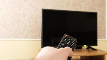 Hand, die eine Fernsehfernbedienung hält und Programme im Fernsehen surft. fernsehen, schalten Sie den Fernseher im Wohnzimmer oder Schlafzimmer auf dem schwarzen Nachttisch ein oder aus. Platz kopieren.