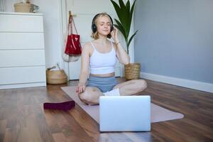Porträt von passen und gesund Frau beim heim, trainieren Yoga, Sitzung auf Gummi Matte, Hören zu Anleitung online, mit Meditation Musik- zu entspannen, folgenden Orientierungshilfe auf Laptop foto
