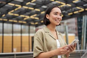 Technologie und Personen. lächelnd asiatisch Mädchen mit Handy, Mobiltelefon Telefon, mit Telefon und Gehen im Stadt im Tageslicht foto