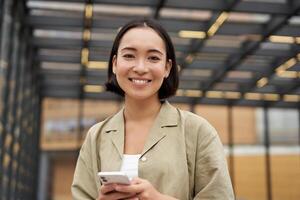 Technologie und Personen. lächelnd asiatisch Mädchen mit Handy, Mobiltelefon Telefon, mit Telefon und Gehen im Stadt im Tageslicht foto