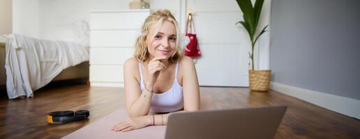 Porträt von jung Fitness Frau, tun trainieren beim heim, suchen beim ihr Fitnessstudio Lehrer auf Laptop Bildschirm, tun online Ausbildung Session foto
