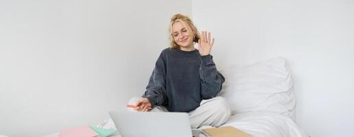 Porträt von Schön, lächelnd jung Frau Sitzung auf Bett, Wellen ihr Hand beim Laptop Kamera, sagt Hallo zu Freund auf Plaudern Anwendung, Studien beim Zuhause auf Fernbedienung, verbindet zu online Kurs foto