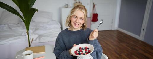 Porträt von lächelnd blond Frau, Essen Frühstück, halten Schüssel und Löffel, Sitzung im Schlafzimmer, suchen glücklich beim Kamera foto