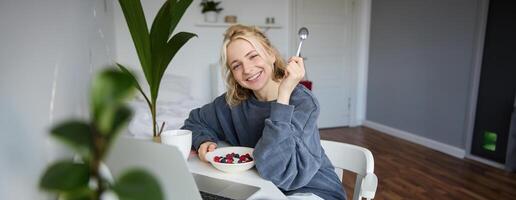 Porträt von lächelnd blond jung Frau, Essen im Vorderseite von Laptop, Aufpassen s online während haben Frühstück, genießen Nachtisch, Sitzung im Schlafzimmer foto