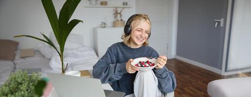 Bild von glücklich Frau Sitzung im ein Zimmer, Aufpassen interessant Fernseher Show oder Film auf Laptop, mit schreiend Service, tragen Kopfhörer, Essen Dessert und Trinken Tee foto