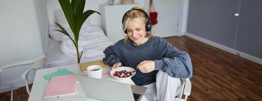 Porträt von glücklich jung blond Frau, Sitzung im ein Zimmer, Aufpassen Film auf Laptop und Essen gesund Frühstück, Trinken Tee, ruhen auf Wochenende foto