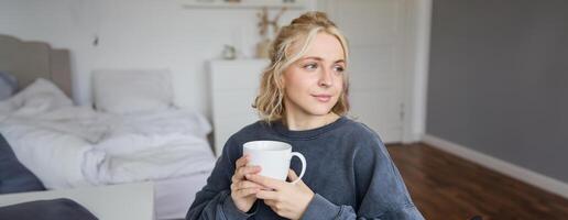 schließen oben Porträt von lächelnd jung blond Frau, sitzt mit Tasse von Tee im Schlafzimmer, ruht beim heim, genießt ihr Kaffee foto