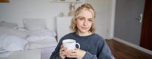 schließen oben Porträt von lächelnd jung blond Frau, sitzt mit Tasse von Tee im Schlafzimmer, ruht beim heim, genießt ihr Kaffee foto