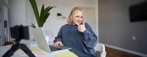 Porträt von lächelnd jung blond Frau, Mädchen Schüler studieren von heim, mit online Akademie, tun Kurs im Internet, mit Laptop, zeigen Daumen hoch. Konzept von Entfernung Lernen foto