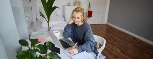 Lebensstil Bild von jung Frau Schreiben Nieder etwas im Notizbuch, Herstellung Anmerkungen, studieren online, tun Kurs im Internet, Hören zu interessant Information, mit Laptop foto