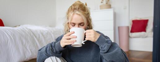 Porträt von jung Frau Sitzung auf Schlafzimmer Boden, Trinken Tee, halten Weiß Becher und lächelnd beim Kamera foto