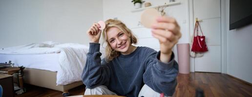 Porträt von jung Frau, Inhalt Schöpfer, zeigen Schönheit bilden Produkte beim Kamera, Sitzung auf Fußboden im Schlafzimmer und lächelnd foto