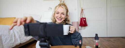 Porträt von süß lächelnd Frau, blond Mädchen Getränke Tee und Aufzeichnungen ein lässig, Lebensstil Blog, vlogger sitzt im ein Zimmer mit Kamera und Stabilisator, hält Tasse von Kaffee und Gespräche foto