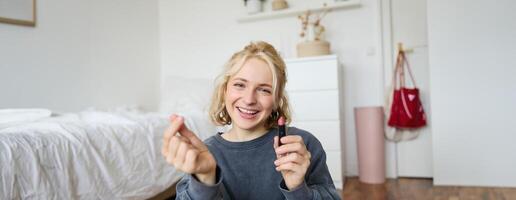 Porträt von jung glücklich Frau, Inhalt Medien Schöpfer Aufzeichnung ein vlog Über bilden im ihr Zimmer, mit Digital Kamera und Stabilisator zu Show Schönheit Produkte, halten Lippenstift foto