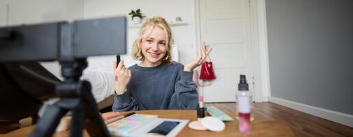 Bild von jung stilvoll Frau, Blogger Aufzeichnung ein Schönheit Lebensstil von ihr pflücken Beste Lippenstift, zeigen Lippe Balsam Farbfelder auf ihr Haut, Sitzung im Vorderseite von Digital Kamera im leeren Zimmer foto