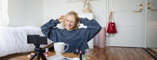 Porträt von süß lächelnd Frau, blond Mädchen Getränke Tee und Aufzeichnungen ein lässig, Lebensstil Blog, vlogger sitzt im ein Zimmer mit Kamera und Stabilisator, hält Tasse von Kaffee und Gespräche foto