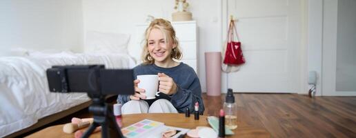 Porträt von süß lächelnd Frau, blond Mädchen Getränke Tee und Aufzeichnungen ein lässig, Lebensstil Blog, vlogger sitzt im ein Zimmer mit Kamera und Stabilisator, hält Tasse von Kaffee und Gespräche foto