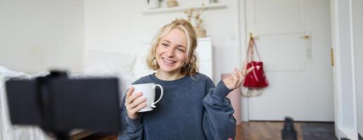 Porträt von süß lächelnd Frau, blond Mädchen Getränke Tee und Aufzeichnungen ein lässig, Lebensstil Blog, vlogger sitzt im ein Zimmer mit Kamera und Stabilisator, hält Tasse von Kaffee und Gespräche foto