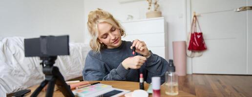 Frau Schönheit Blogger, Aufzeichnungen von Sie selber Sitzung im ein Zimmer und Bewertung bilden Produkte, setzt auf machen hoch, hält Lippenstift und kosmetisch Bürste im Hand, mit Fachmann Kamera zum Inhalt Schaffung foto