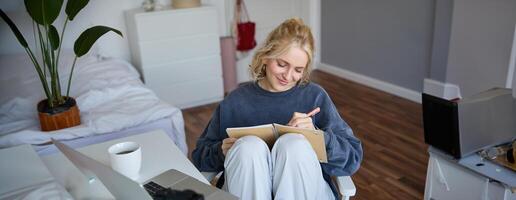 Porträt von jung Frau Schreiben im Tagebuch, Herstellung Anmerkungen im Notizbuch, Sitzung auf Stuhl im ein Zimmer im Vorderseite von Laptop, Schüler tun Hausaufgaben foto