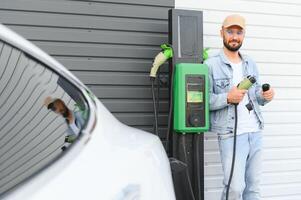 Mann halten Leistung Laden Kabel zum elektrisch Auto im draussen Auto Park. und er s gehen zu verbinden das Auto zu das Laden Bahnhof im das Parkplatz Menge in der Nähe von das Einkaufen Center. foto