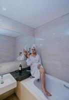asiatisch Frau Sitzung auf Bad oben Innerhalb Hotel Badezimmer tragen Bademantel, Brille und halten Glas von rot Wein. foto