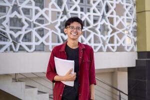 asiatisch Hochschule Schüler lächelnd und suchen. Porträt von ein Hochschule Schüler beim Campus. asiatisch männlich Schüler Stehen halten ein Buch suchen beim das Kamera. foto