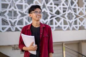 asiatisch Hochschule Schüler lächelnd und suchen. Porträt von ein Hochschule Schüler beim Campus. asiatisch männlich Schüler Stehen halten ein Buch suchen beim das Kamera. foto