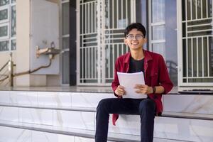 asiatisch Hochschule Schüler lächelnd und suchen. Porträt von ein Hochschule Schüler beim Campus. asiatisch männlich Schüler Sitzung halten ein Buch suchen beim das Kamera. foto