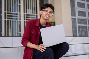 Porträt von asiatisch Hochschule Schüler mit Laptop suchen zu Kamera. ein Mann Arbeiten mit ein Laptop Computer beim Campus foto