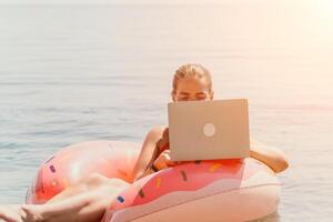 Frau Freiberufler funktioniert auf Laptop Schwimmen im Meer auf Rosa aufblasbar Ring. glücklich Tourist schwebend auf aufblasbar Krapfen und Arbeiten auf Laptop Computer im Ruhe Ozean. freiberuflich, Fernbedienung Arbeiten irgendwo foto