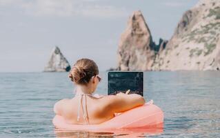 Frau Freiberufler funktioniert auf Laptop Schwimmen im Meer auf Rosa aufblasbar Ring. glücklich Tourist im Sonnenbrille schwebend auf aufblasbar Krapfen und Arbeiten auf Laptop Computer im Ruhe Ozean. Fernbedienung Arbeiten irgendwo foto