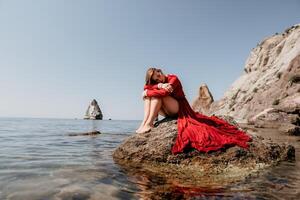 Frau Reise Meer. glücklich Tourist im rot Kleid genießen nehmen Bild draußen zum Erinnerungen. Frau Reisender posieren auf das Felsen beim Meer Bucht umgeben durch vulkanisch Berge, Teilen Reise Abenteuer Reise foto