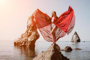 Frau Reise Meer. jung glücklich Frau im ein lange rot Kleid posieren auf ein Strand in der Nähe von das Meer auf Hintergrund von vulkanisch Felsen, mögen im Island, Teilen Reise Abenteuer Reise foto