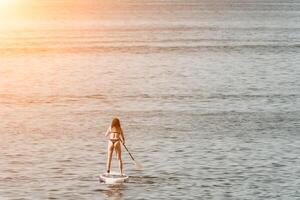 Meer Frau sup. Silhouette von glücklich positiv jung Frau im Bikini, Surfen auf sup Planke, zuversichtlich Paddeln durch Wasser Oberfläche. idyllisch Sonnenuntergang. aktiv Lebensstil beim Meer oder Fluss. schleppend Bewegung. foto