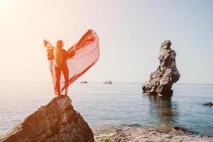 Frau Reise Meer. jung glücklich Frau im ein lange rot Kleid posieren auf ein Strand in der Nähe von das Meer auf Hintergrund von vulkanisch Felsen, mögen im Island, Teilen Reise Abenteuer Reise foto