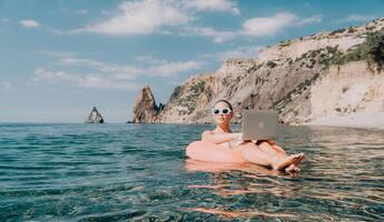 Frau Freiberufler funktioniert auf Laptop Schwimmen im Meer auf Rosa aufblasbar Ring. glücklich Tourist im Sonnenbrille schwebend auf aufblasbar Krapfen und Arbeiten auf Laptop Computer im Ruhe Ozean. Fernbedienung Arbeiten irgendwo foto
