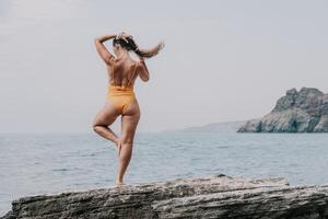 Frau Meer Yoga. zurück Aussicht von kostenlos Ruhe Glückseligkeit zufrieden Frau mit lange Haar Stehen auf oben Felsen mit Yoga Position gegen von Himmel durch das Meer. gesund Lebensstil draußen im Natur, Fitness Konzept. foto