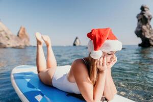 Frau Meer sup. schließen oben Porträt von glücklich jung kaukasisch Frau mit lange Haar im Santa Hut suchen beim Kamera und lächelnd. süß Frau Porträt im ein Weiß Bikini posieren auf sup Tafel im das Meer foto