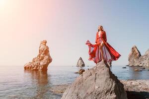 Frau Reise Meer. jung glücklich Frau im ein lange rot Kleid posieren auf ein Strand in der Nähe von das Meer auf Hintergrund von vulkanisch Felsen, mögen im Island, Teilen Reise Abenteuer Reise foto