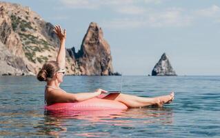 Frau Freiberufler funktioniert auf Laptop Schwimmen im Meer auf Rosa aufblasbar Ring. glücklich Tourist im Sonnenbrille schwebend auf aufblasbar Krapfen und Arbeiten auf Laptop Computer im Ruhe Ozean. Fernbedienung Arbeiten irgendwo foto