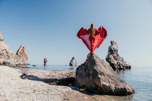 Frau Reise Meer. jung glücklich Frau im ein lange rot Kleid posieren auf ein Strand in der Nähe von das Meer auf Hintergrund von vulkanisch Felsen, mögen im Island, Teilen Reise Abenteuer Reise foto